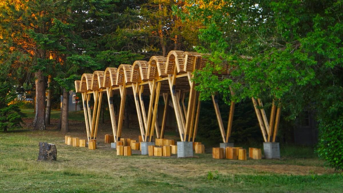 A rolling roof and V-shaped framed outdoor classroom sits before trees.